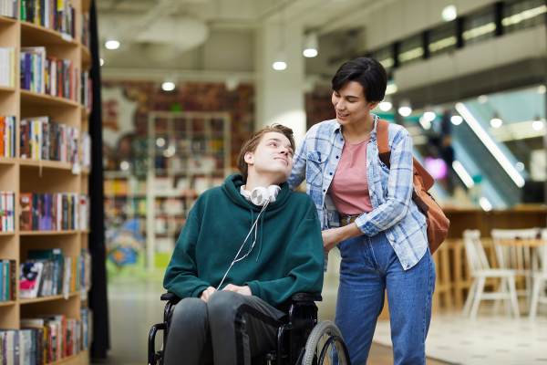 Eine Betreuerin schiebt einen behinderten Menschen im Rollstuhl durch eine Bücherei. Beide lächeln